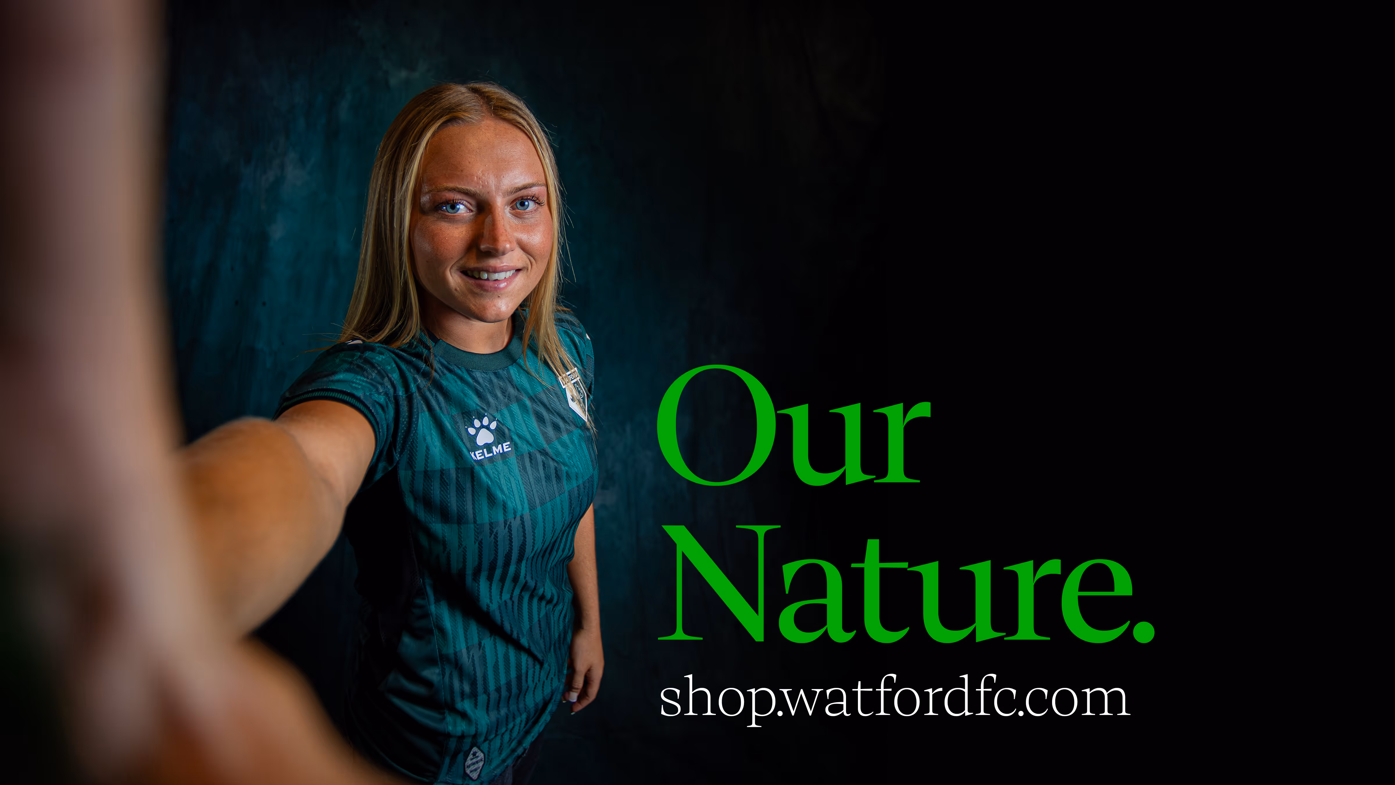 Football player, Maddie Perry, in green Watford FC jersey with hand raised towards camera with text ‘Our Nature’ and shop.watfordfc.com on dark background.