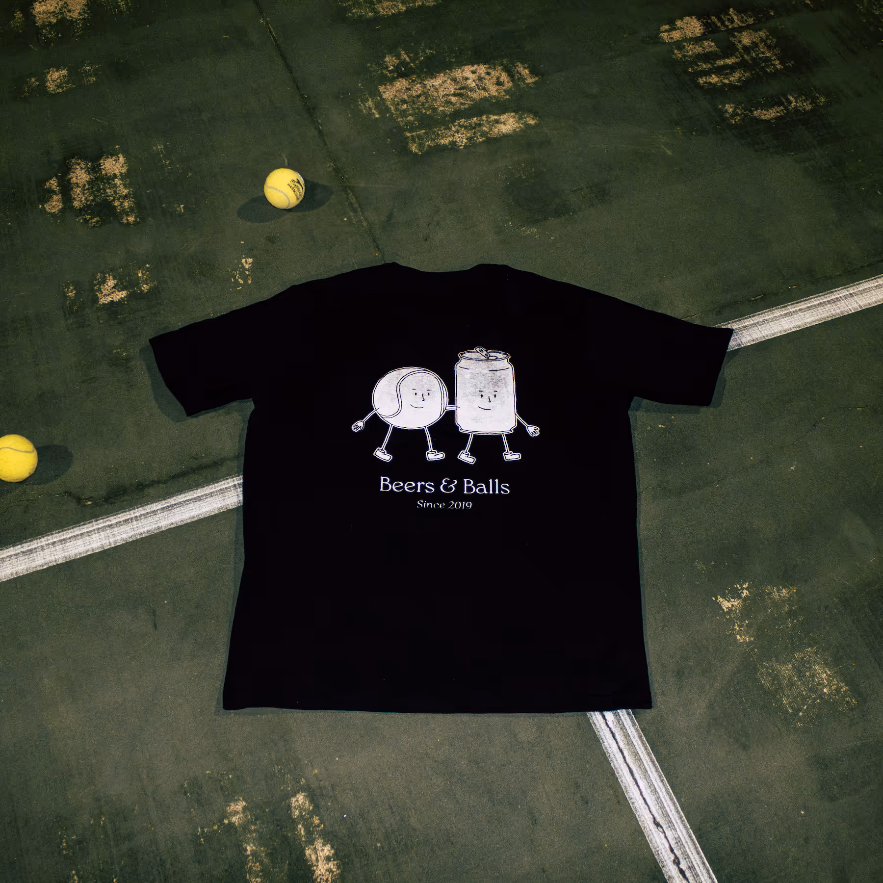 Black t-shirt laid on a tennis court with a cartoon tennis ball and beer can holding each other by the shoulders and the text 'Beers & Balls Since 2019'.