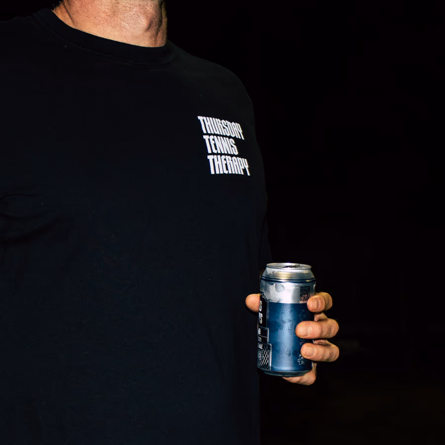 Person wearing a black shirt with text 'Thursday Tennis Therapy' holding a blue can.