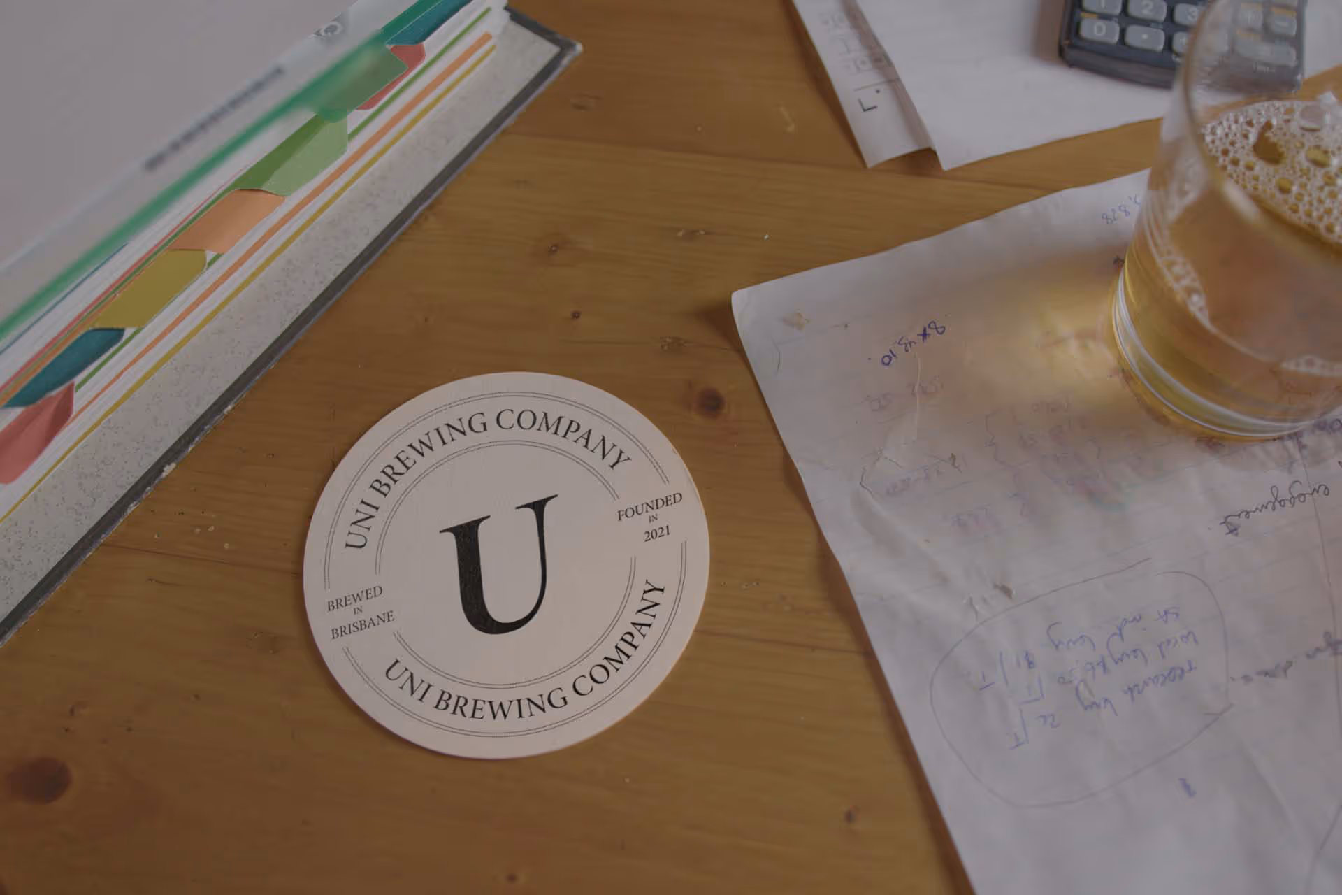 Wooden desk with a round Uni Brewing Company coaster, a partially filled beer glass, scattered papers with handwritten notes, and a colorful ring binder.