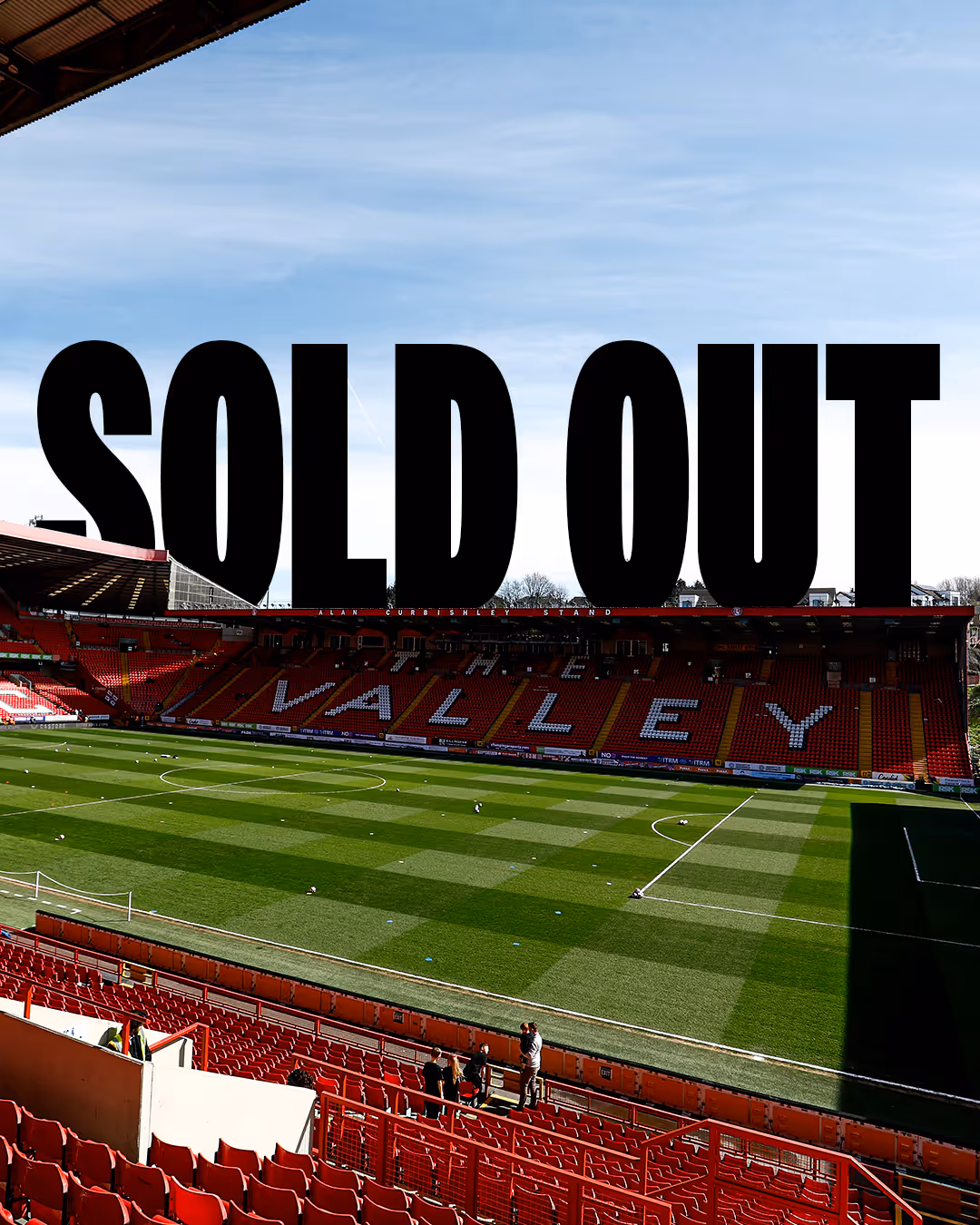A shot of The Valley, Charlton Athletic's football stadium with red seats and green field, with large black text reading 'Sold Out' in the sky above the stands.