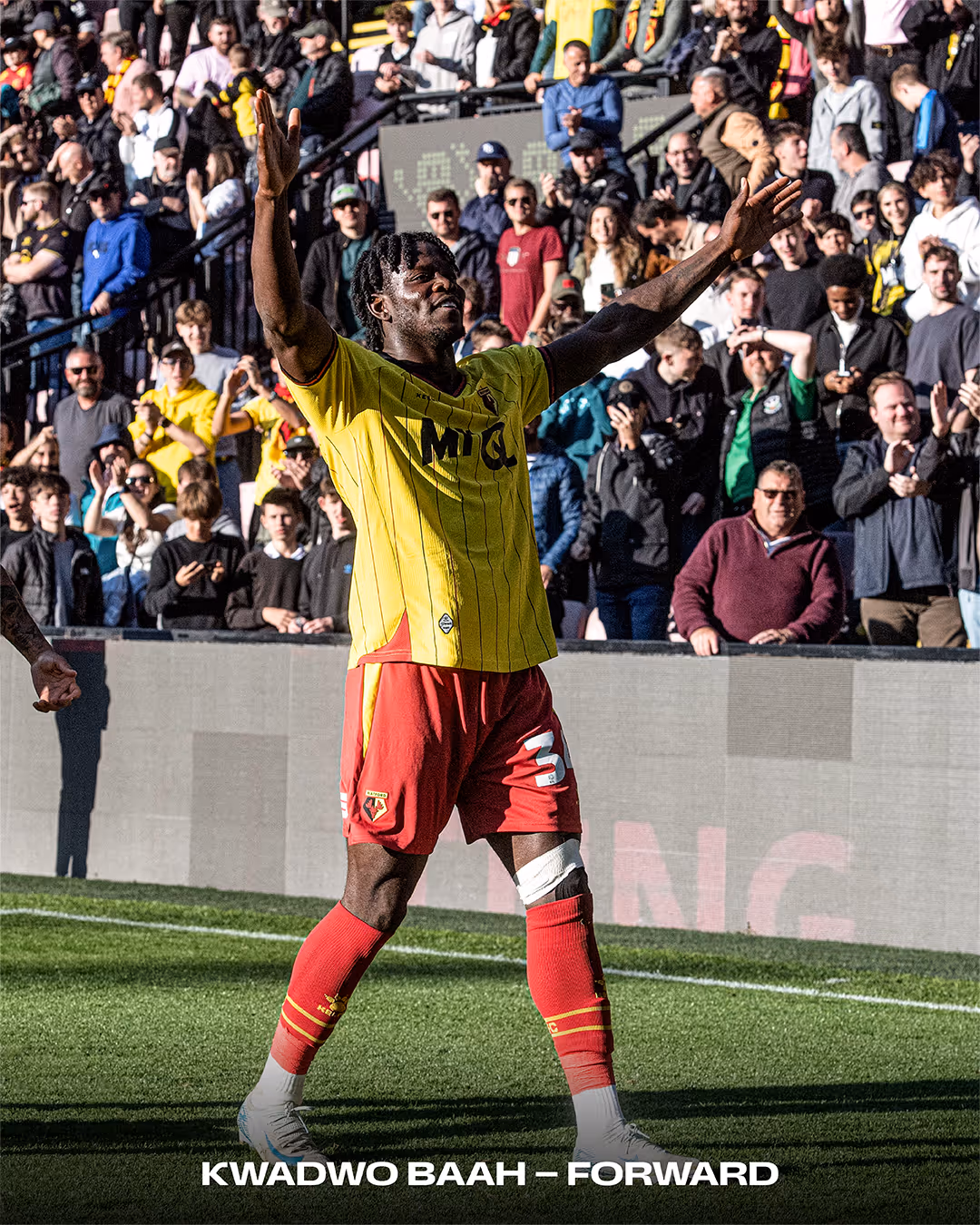 Kwadwo Baah celebrating on a football field wearing a yellow and red Watford FC kit with fans clapping in the background.