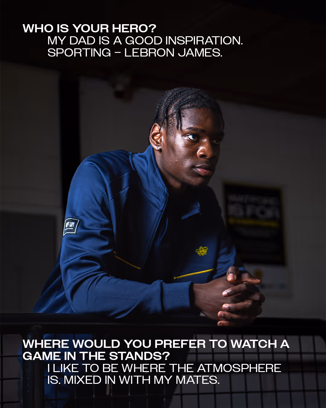 Ian Kamga in a navy blue jacket leaning on a railing inside a dimly-lit Vicarage Road stadium with text about sports heroes and game preferences.