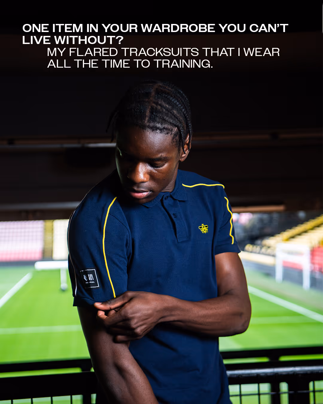 Ian Kamga in a navy blue polo shirt with yellow detailing adjusts his sleeve standing inside Vicarage Road stadium.