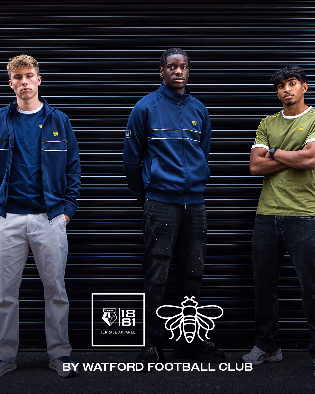 Three Watford academy players standing in front of a black shutter wearing Watford Football Club Terrace Apparel, two in navy blue jackets and one in a khaki green t-shirt.