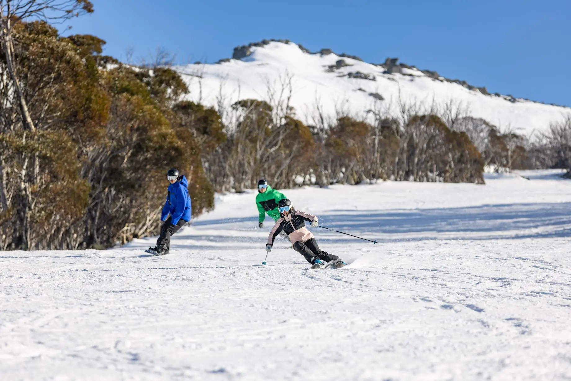 Two skiers on a snowy slope with a clear blue sky and mountain ridge in the background.