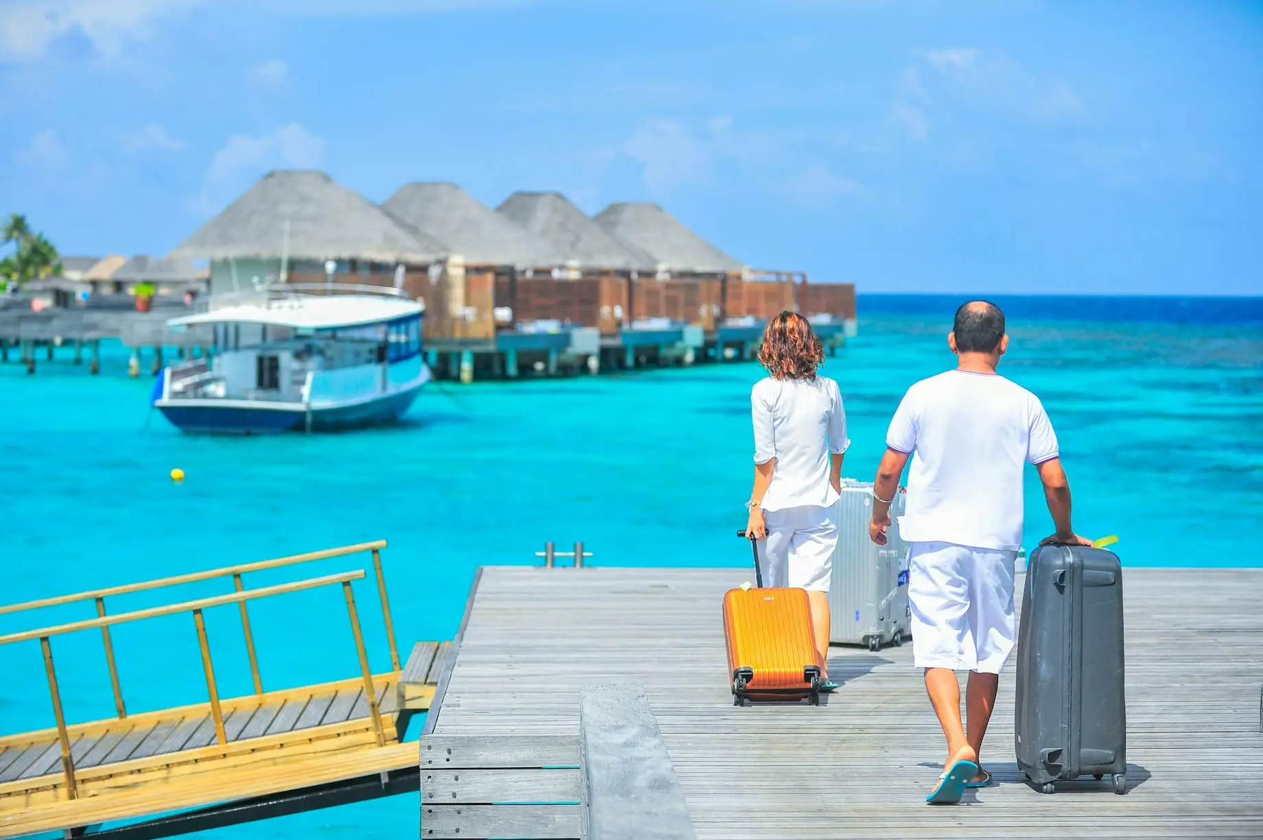 A couple with suitcases walking on a pier towards a boat, with turquoise water and bungalows in the background.