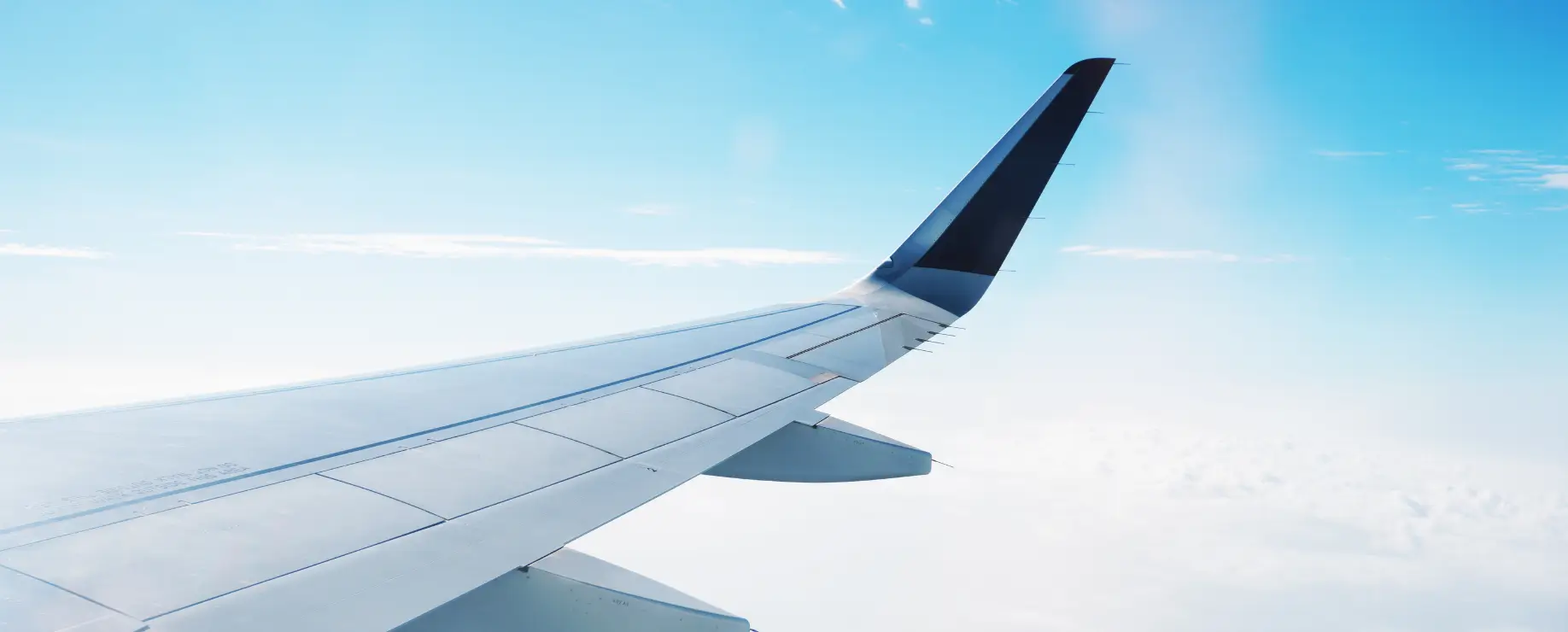 The image shows the wing of an airplane flying above the clouds with a clear blue sky in the background.