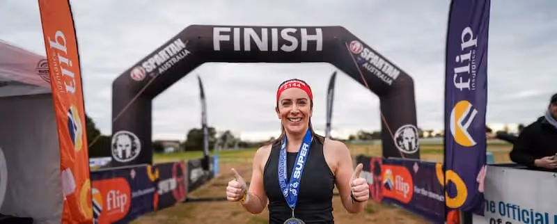 Smiling female Spartan race finisher giving thumbs up at event finish line with Flip Insurance branding in background