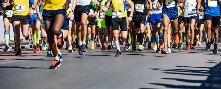 A large group of runners participating in a marathon race, wearing athletic gear and numbered bibs, sprinting forward on a sunlit street.