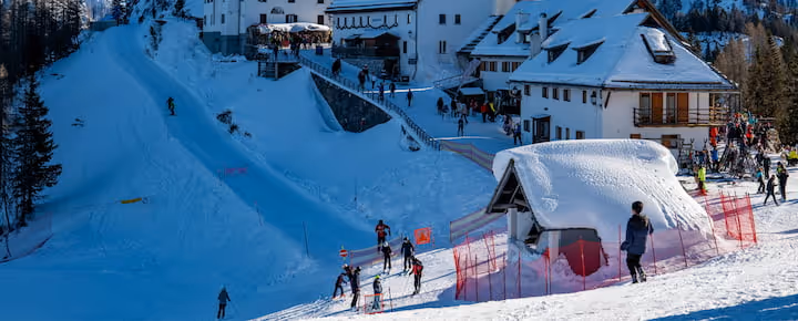 Skiers on a snowy alpine slope with a mountain lodge and red course markers.