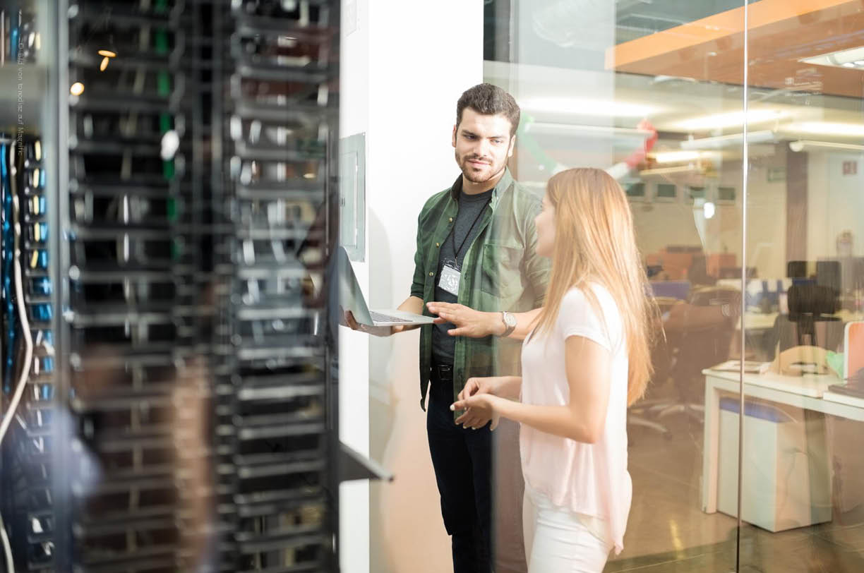 Zwei IT-Fachleute arbeiten konzentriert an einem Laptop, während sie vor einem offenen Serverschrank stehen, der wichtige IT-Systeme und Hardware für die Unternehmens-IT enthält. Sie diskutieren über die IT-Infrastruktur und die Dokumentation, um die Effizienz und Sicherheit in ihrer IT-Umgebung zu verbessern.