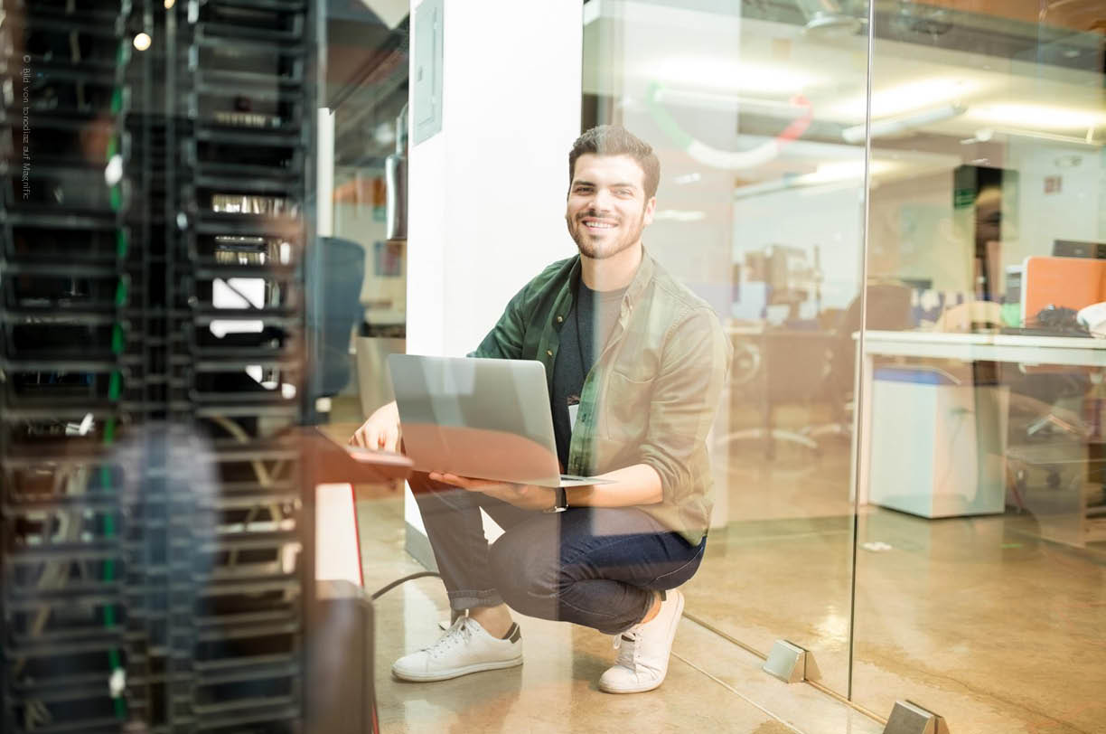 Eine Person hockt vor einem Serverschrank, um wichtige Informationen zur IT-Infrastruktur und zur professionellen IT-Dokumentation zuzugreifen. Der Serverschrank steht in einer modernen IT-Umgebung, die für den Betrieb und das Management von IT-Systemen eingerichtet ist.