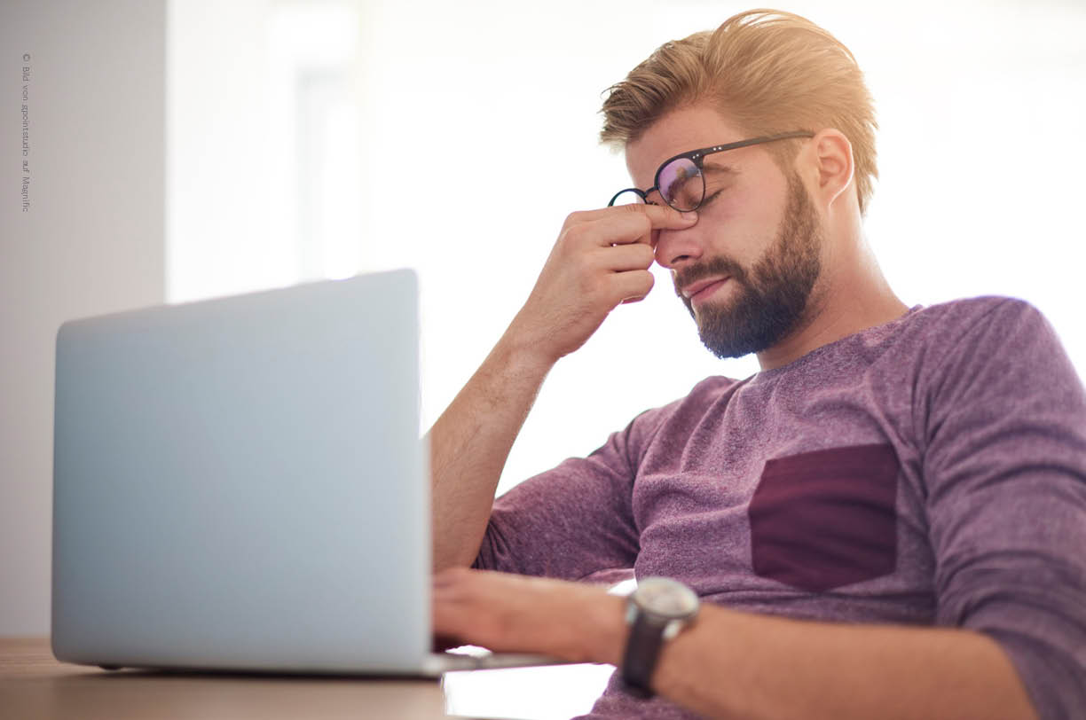 Ein Mitarbeiter sitzt an einem Laptop und überprüft eine verdächtige E-Mail, die möglicherweise Teil einer Phishing-Kampagne ist. Er hält sich vor Verzweifllung die Nase und hat die Augen geschlossen.