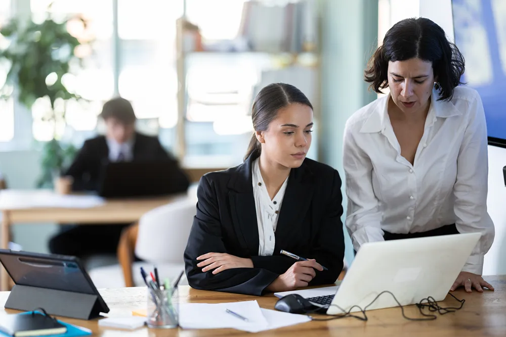 Colleague helping a coworker learn new project management software on a laptop in a modern office