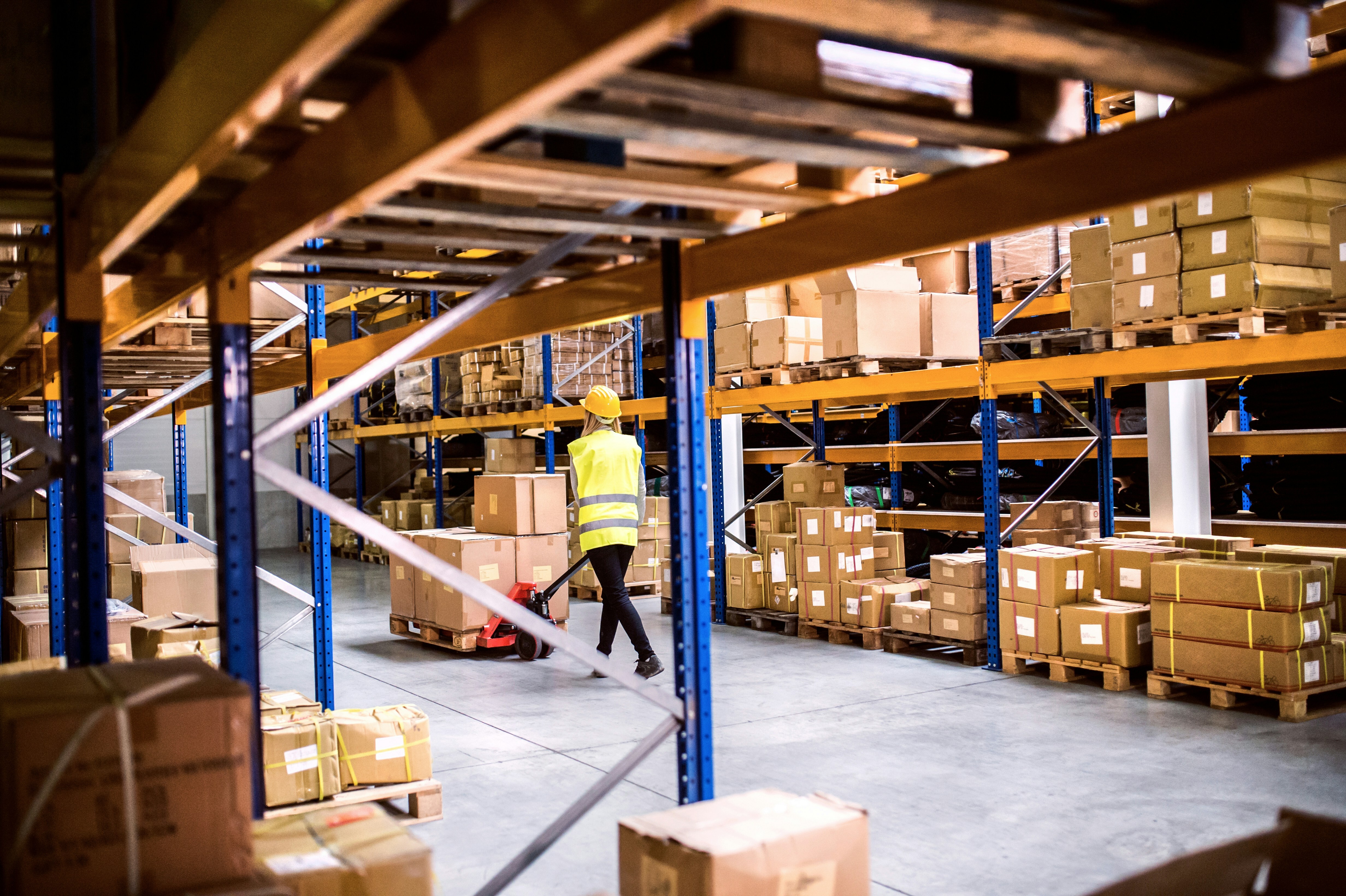 Warehouse worker in yellow safety vest and hard hat pulling a pallet jack through a CPG inventory storage facility