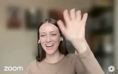 Smiling woman wearing earbuds waving her raised hand during a Zoom video call.