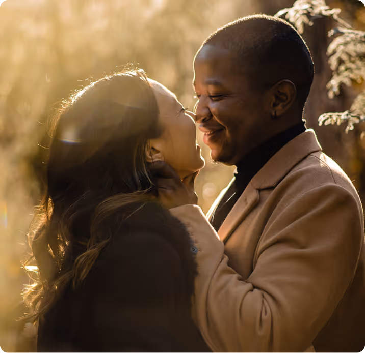 Couple smiling closely with foreheads nearly touching, bathed in warm golden sunlight outdoors.