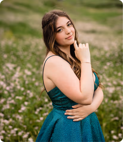 Young woman with long brown hair wearing a sleeveless teal dress standing in a field of green grass and small flowers, looking at the camera with a slight smile.