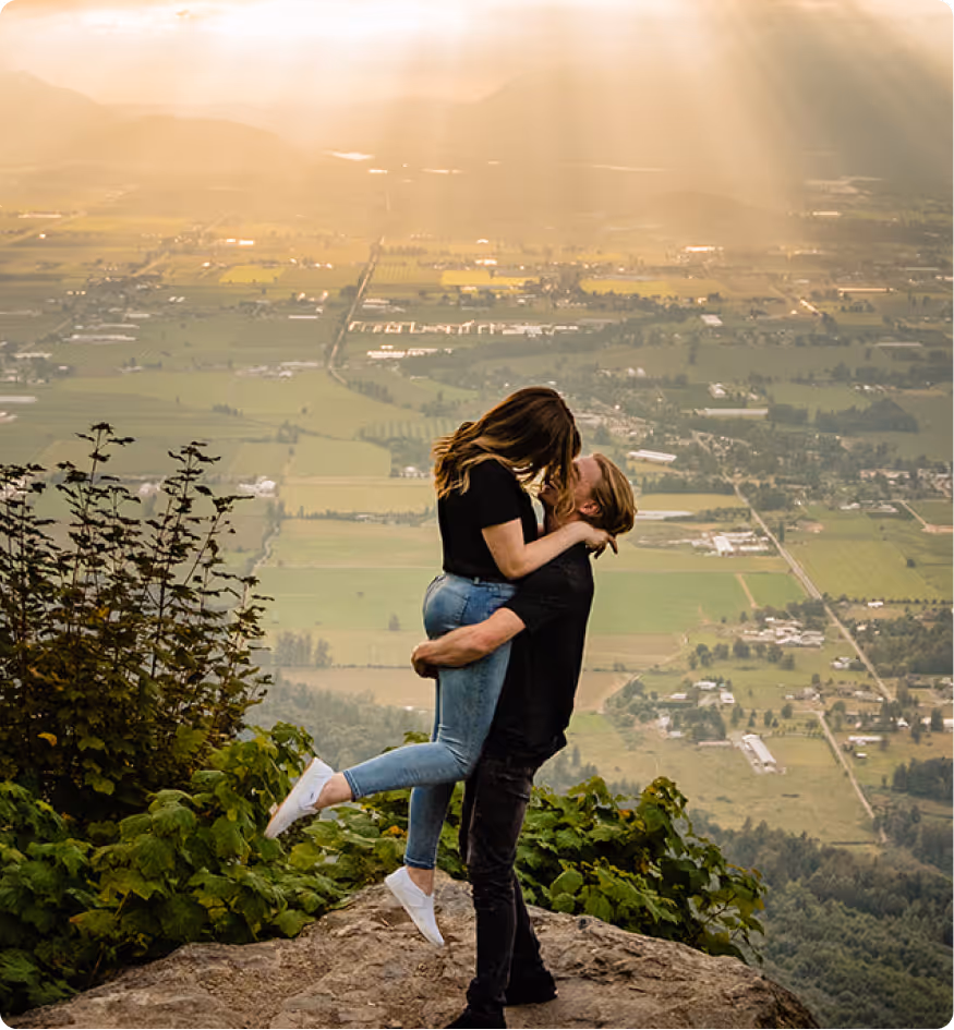 Couple embracing on a cliff overlooking a sunlit valley with patchwork fields and distant houses.