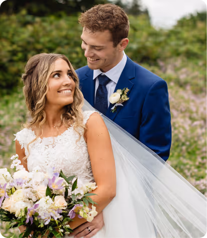 Bride in white lace dress holding a bouquet, smiling at groom in blue suit with boutonniere, outdoors with greenery background.