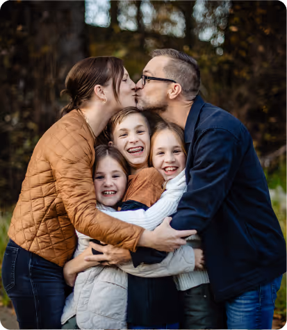 Smiling family of two adults and three children embracing outdoors, with the adults kissing above the children.