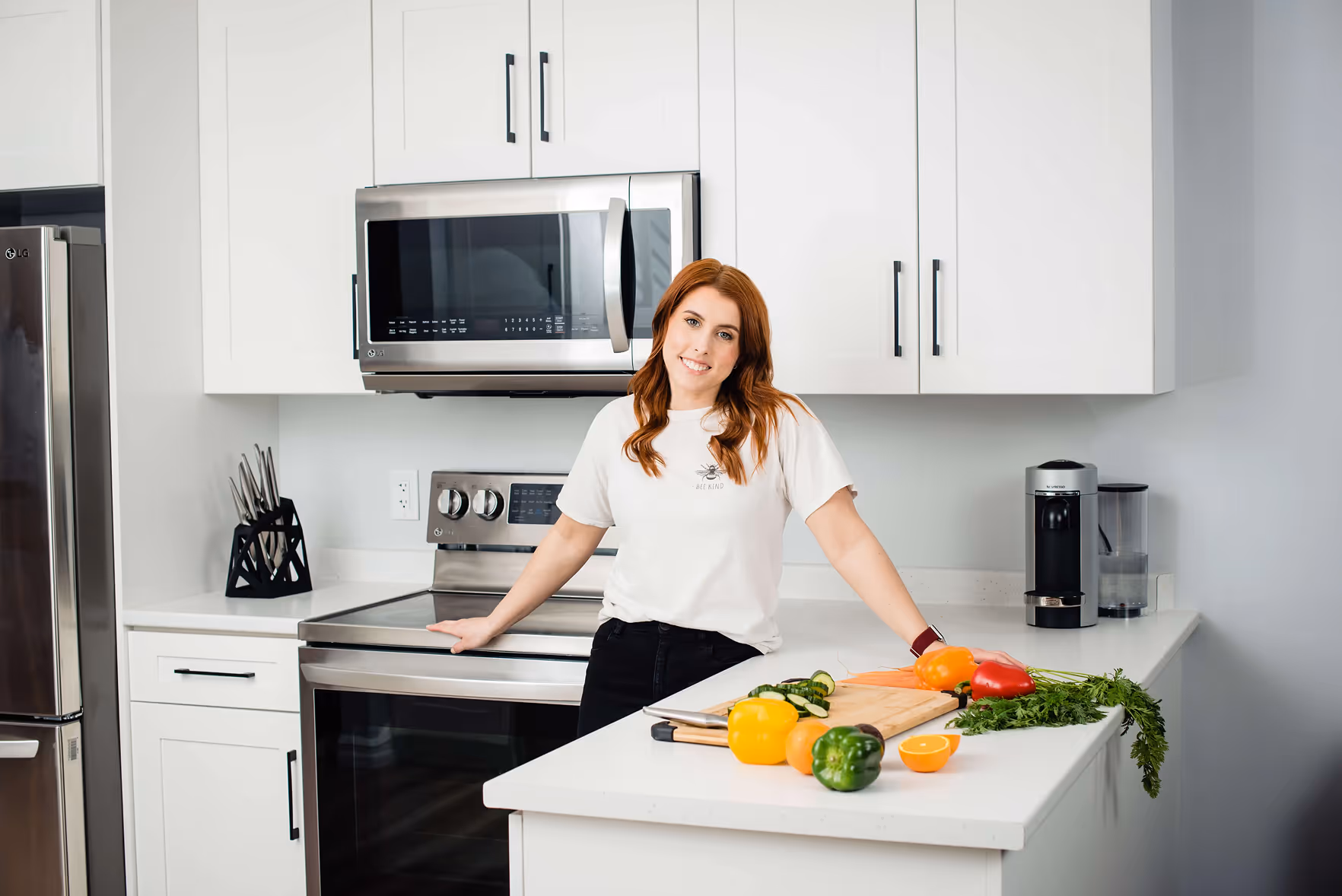 A woman with red hair leaning on a kitchen counter with a cutting board and assorted fresh vegetables including bell peppers, carrots, and an orange half.
