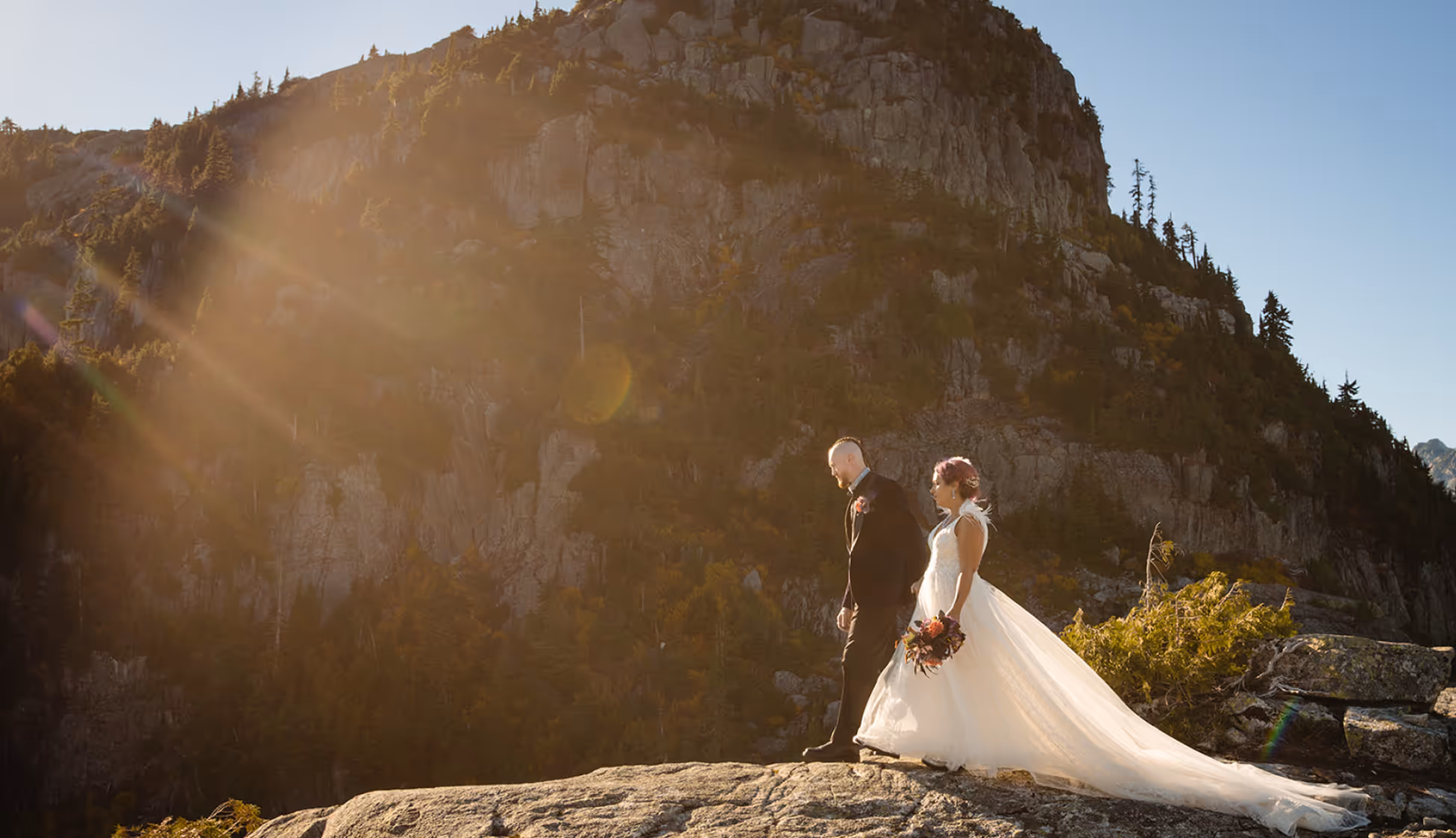 Bride and groom walking on a rocky ledge with a mountainous background bathed in warm sunlight.