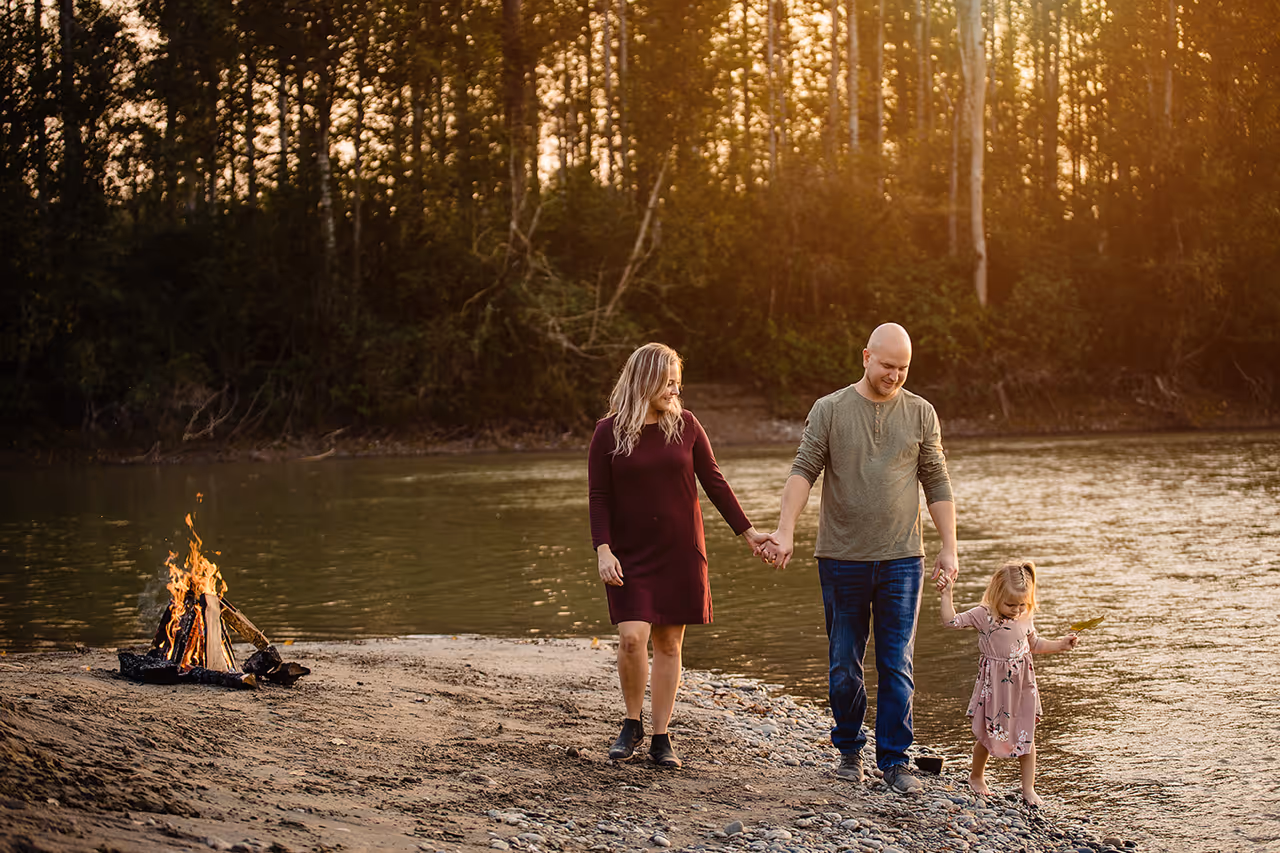 Family of three holding hands while walking barefoot on a riverbank at sunset with a small campfire nearby.