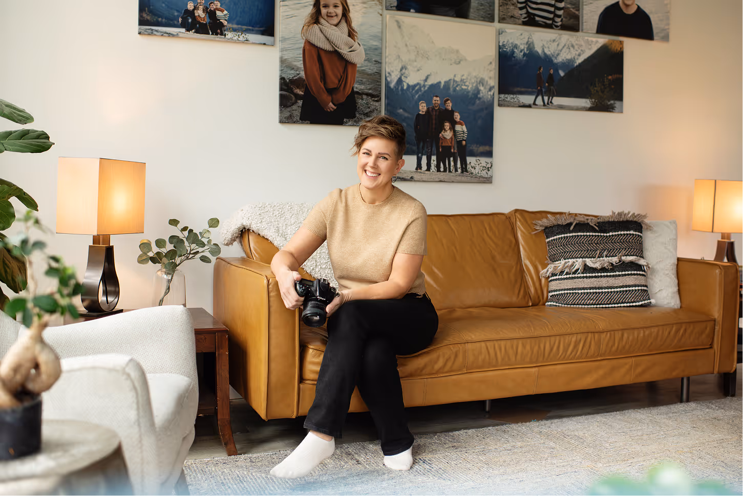 Smiling woman sitting barefoot on a brown leather sofa holding a camera in a cozy living room with family photos on the wall.