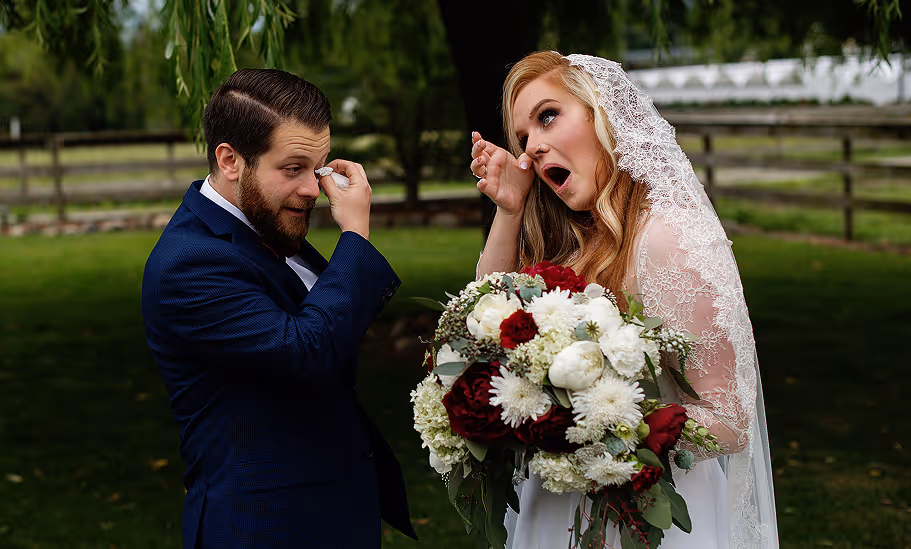 Bride and groom wiping tears of joy during a wedding outdoors with the bride holding a bouquet of red and white flowers.