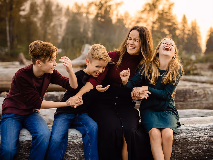 A woman and three children sitting on wooden logs outdoors, laughing and playfully interacting with each other.