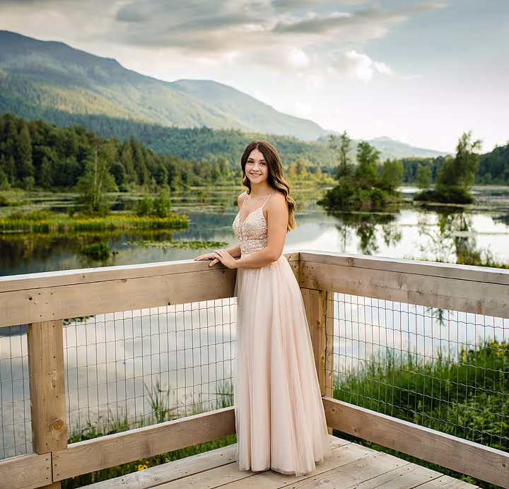 Young woman in a long beige dress standing on a wooden deck overlooking a scenic lake with lush green trees and mountains in the background.