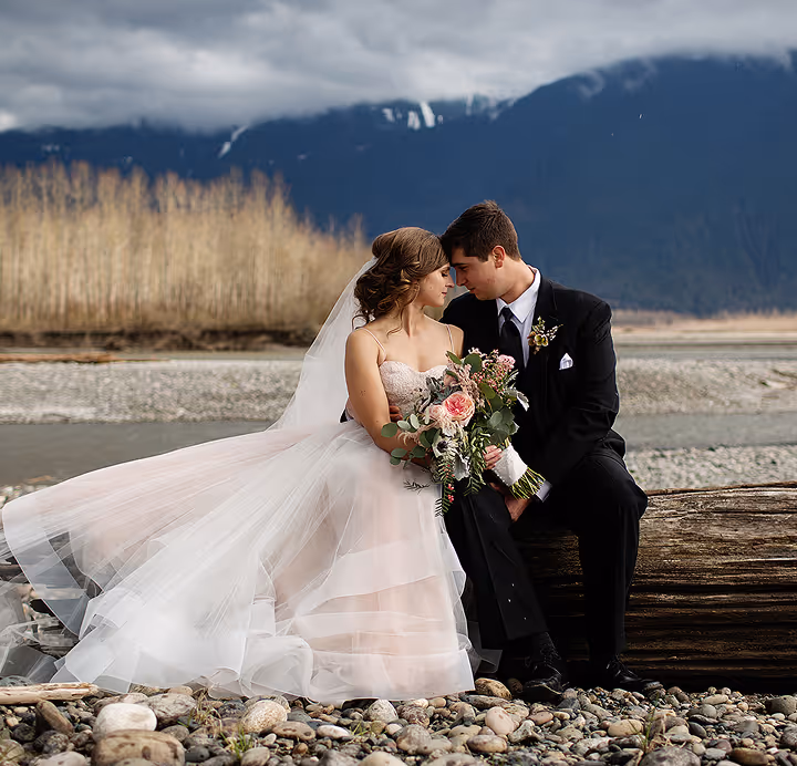 Bride and groom sitting on a log by a rocky riverbank with mountains and cloudy sky in the background, the bride holding a bouquet of flowers.