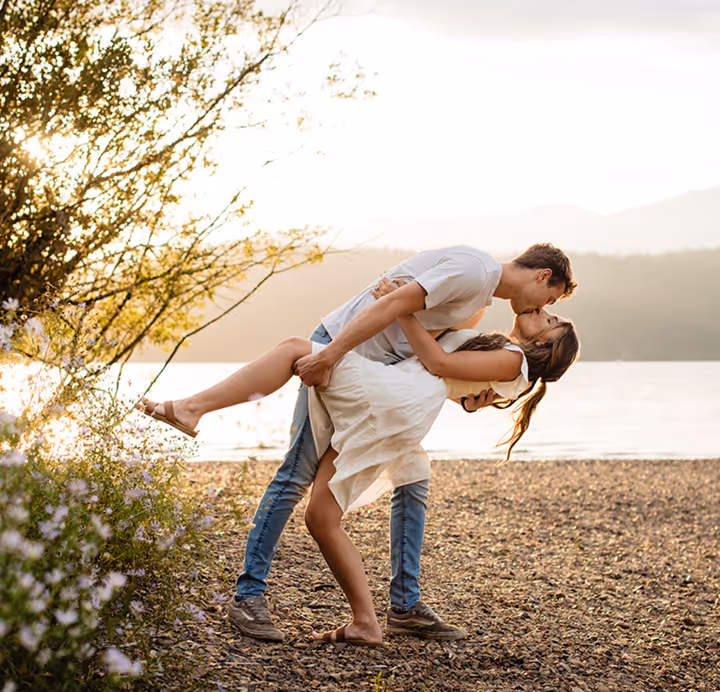 Couple kissing on a rocky beach at sunset, with the man dipping the woman backward near flowering bushes.