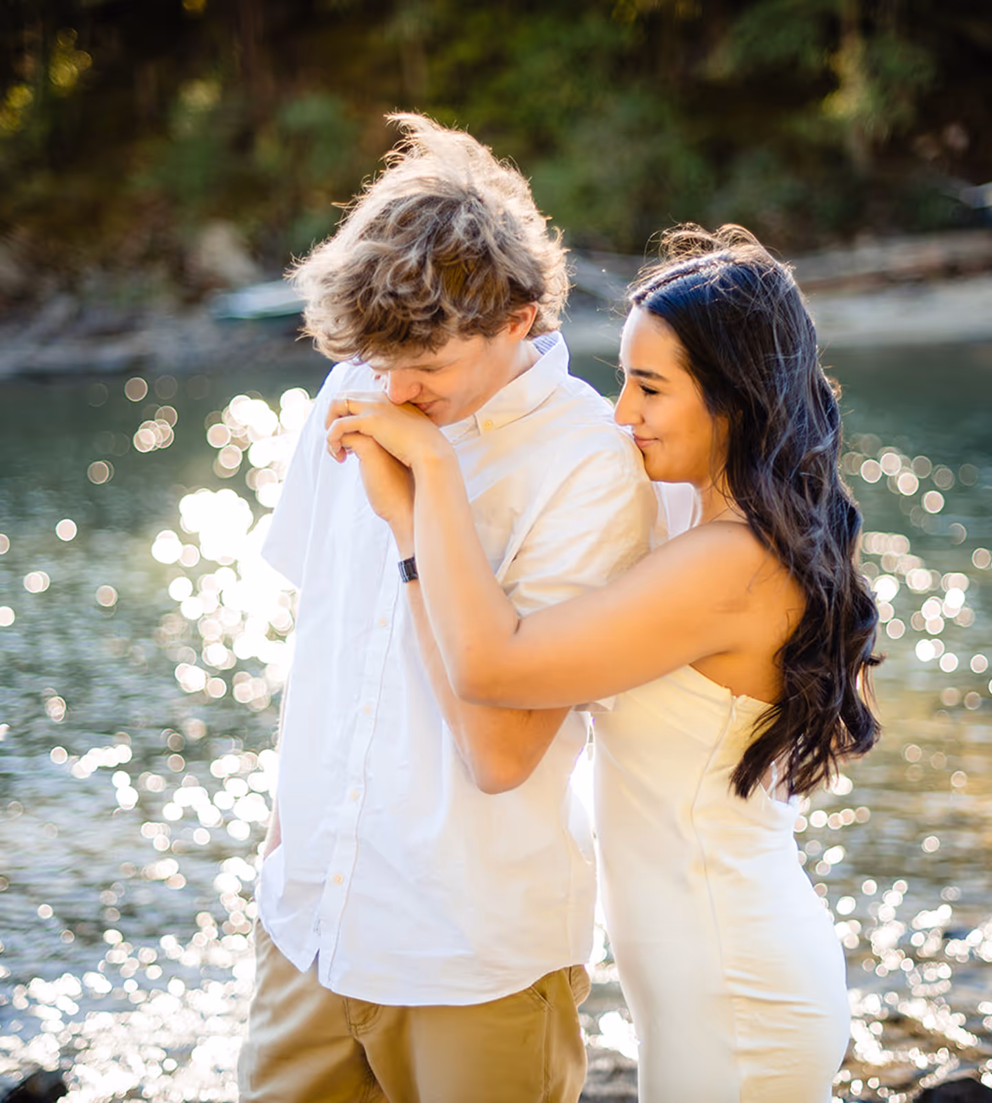 Couple standing by water with woman in white dress embracing man in white shirt who is kissing her hand.