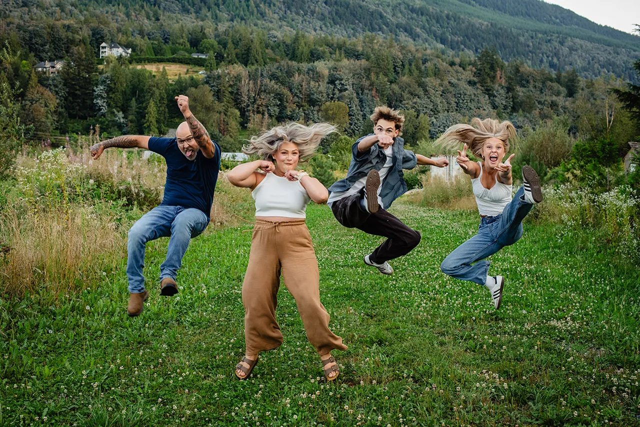 Four people jumping energetically on a green grassy field with forested hills in the background.