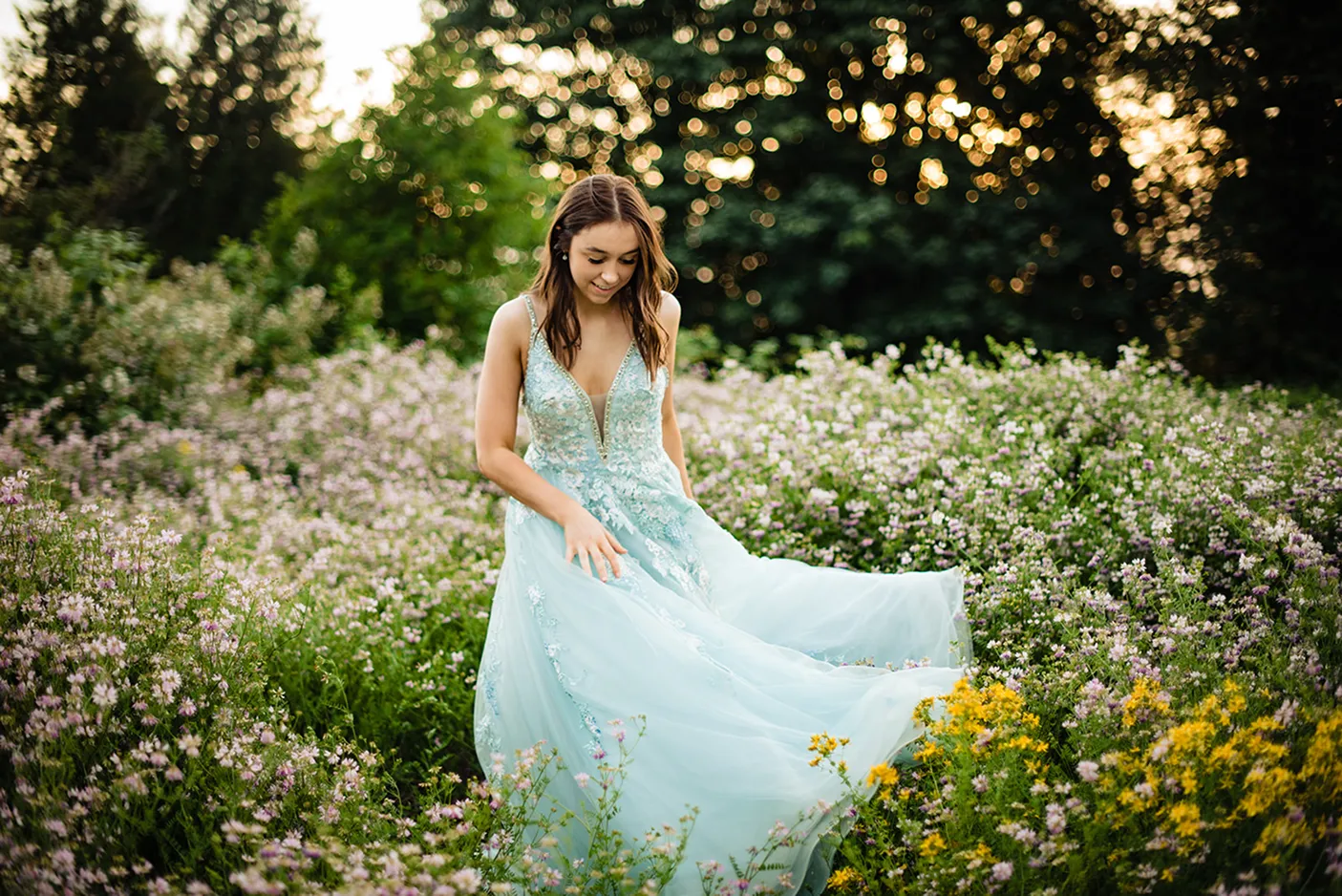 Young woman in a flowing light blue gown standing amidst a field of wildflowers with trees and bokeh sunlight in the background.