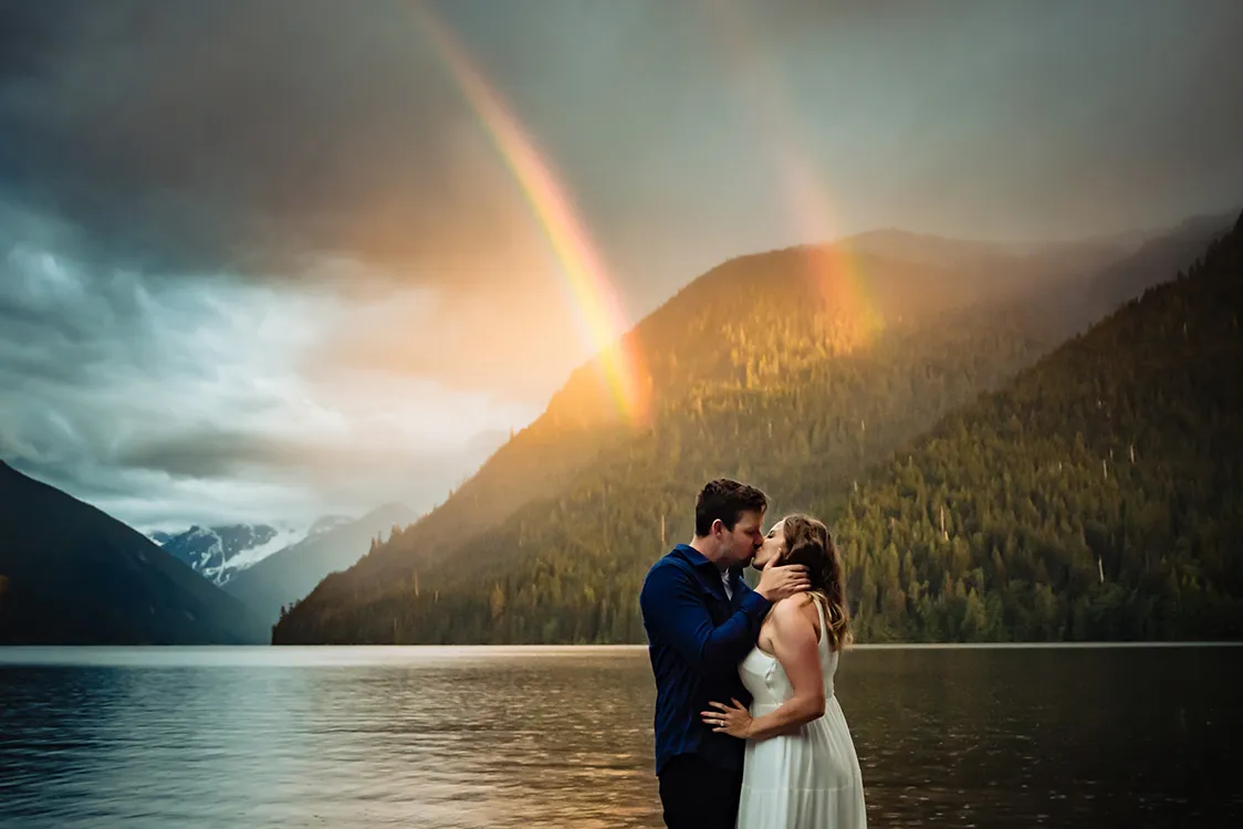 Couple kissing by a lake with forested mountains and a double rainbow in the background.
