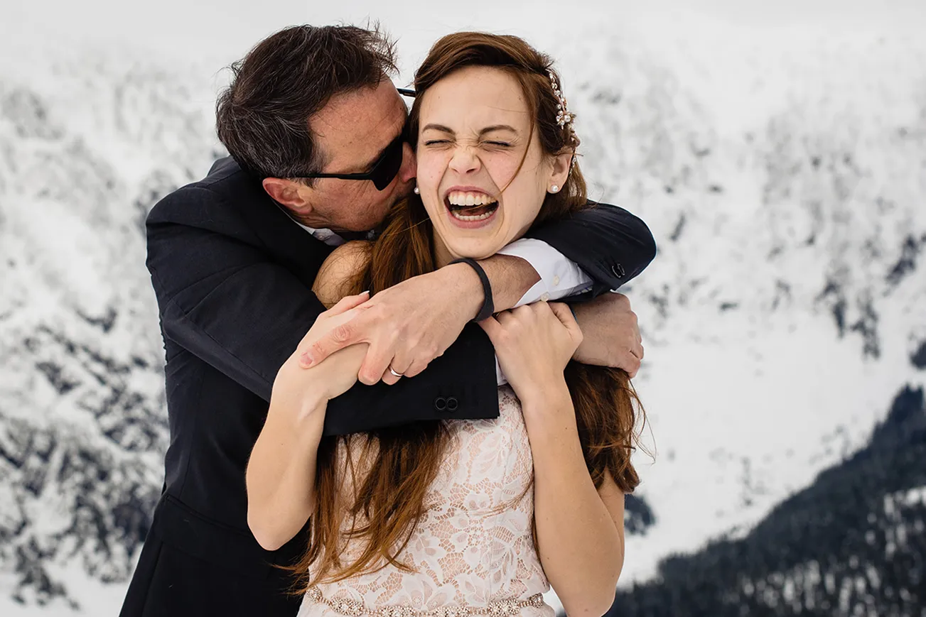 Man in suit embracing and kissing laughing woman in white lace dress with snowy mountain background.