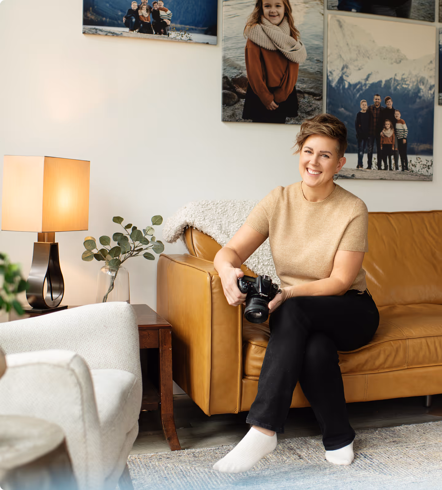 Smiling woman sitting barefoot on a tan leather couch holding a camera in a cozy, well-lit living room.