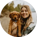 Smiling woman holding a brown puppy outdoors with trees and water in the background.