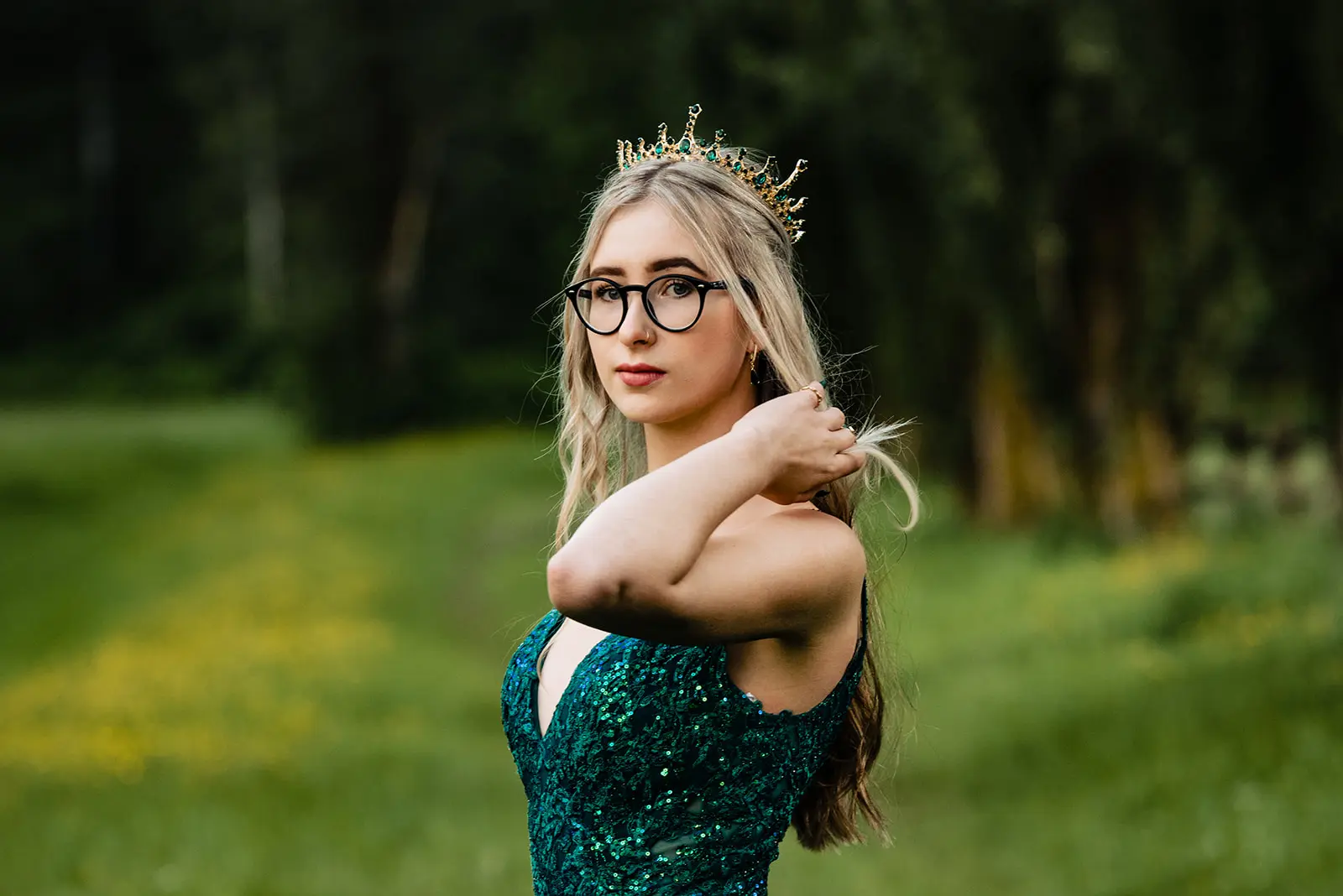 Young woman wearing glasses, a green sequin dress, and a jeweled crown in an outdoor green setting.