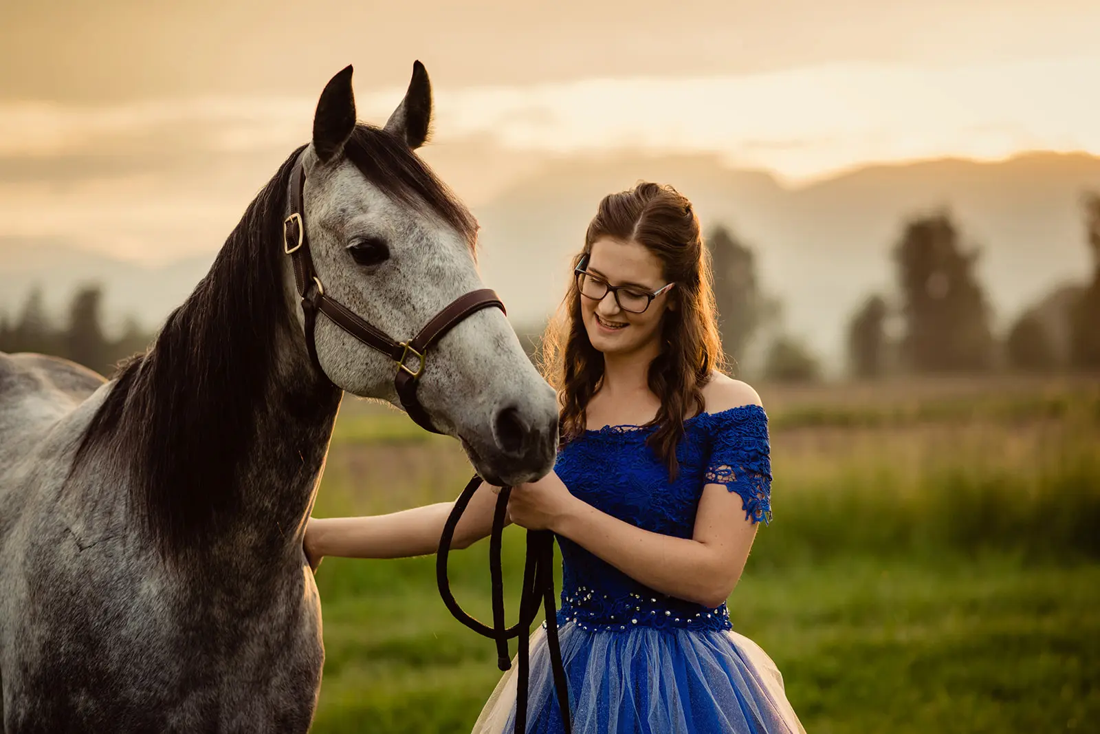 Young woman in blue lace dress smiling while holding a gray horse by the reins outdoors at sunset.