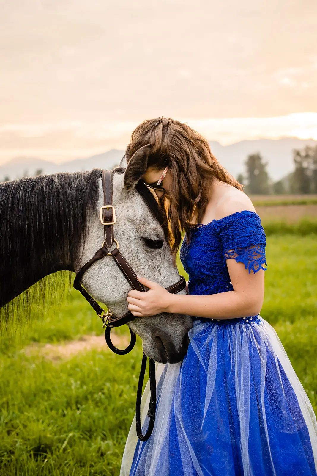 Young woman in a blue dress gently resting her forehead against a gray horse in a green field.