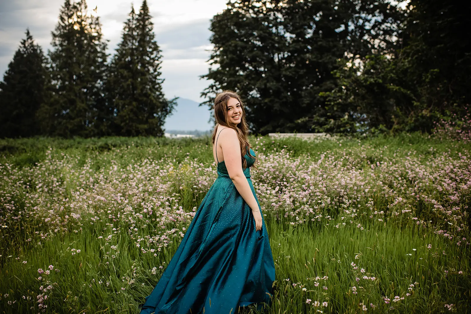 Young woman in a teal strapless gown smiling in a green field with pink wildflowers and trees.