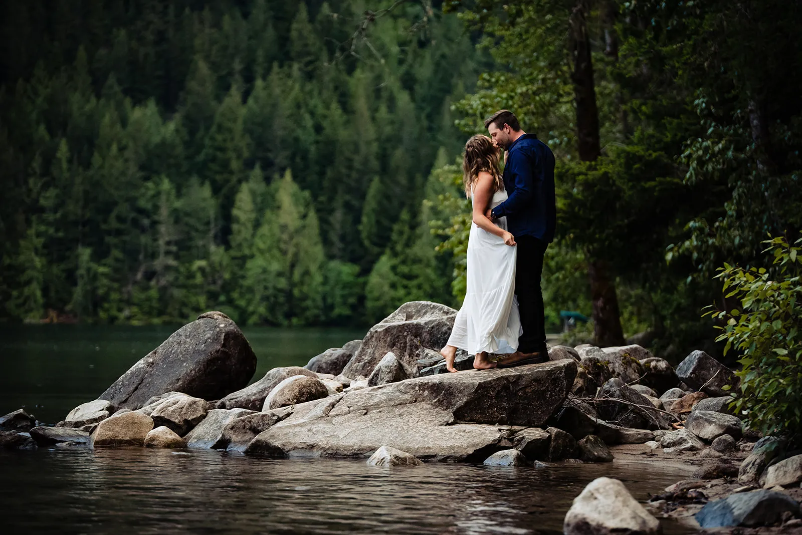 Couple embracing on large rocks by a calm lake with dense forest in the background.