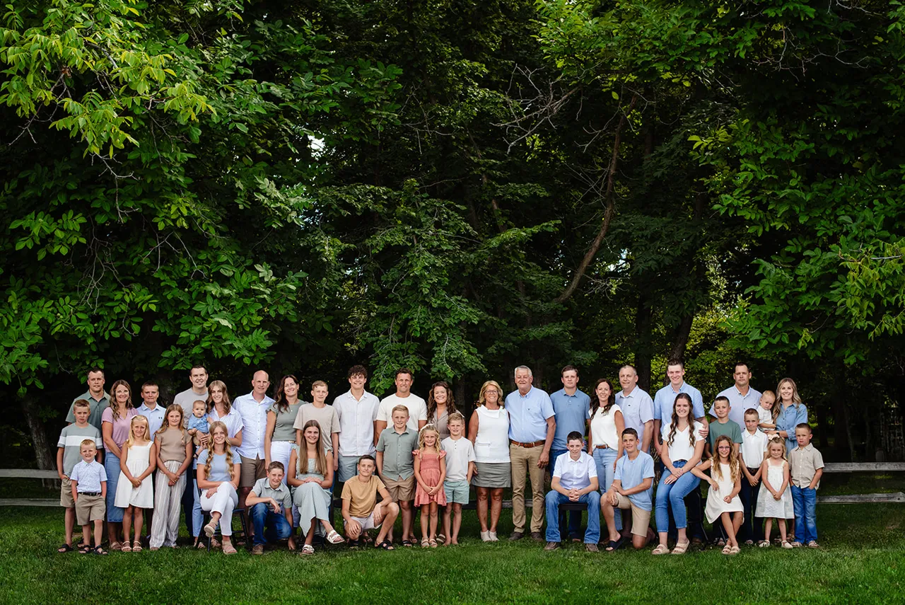 Large multi-generation family posing outdoors on grass with trees in the background.