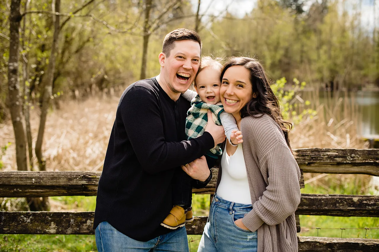 Smiling man and woman holding a happy baby outdoors near a wooden fence with trees in background.