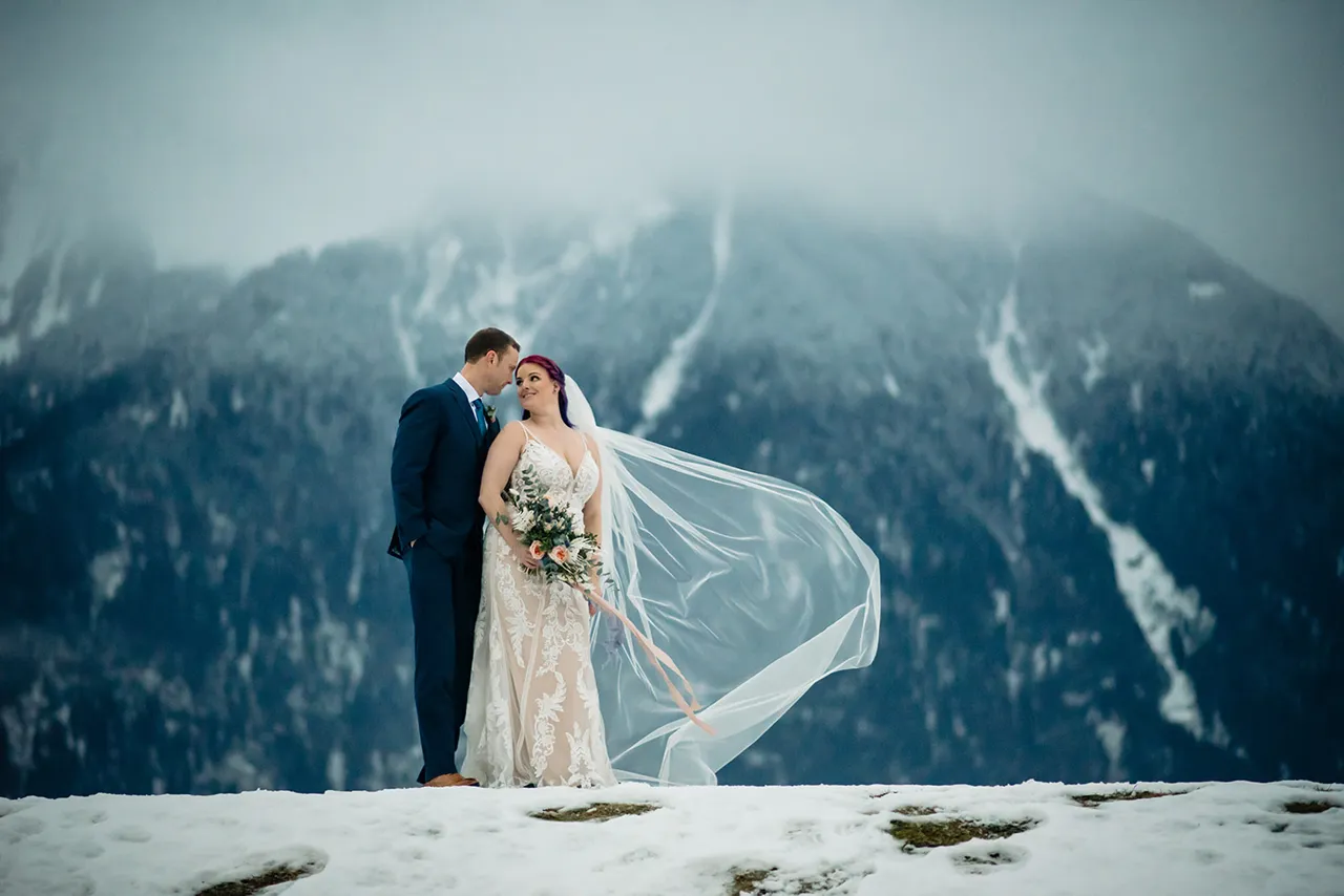 Bride in lace dress and groom in suit standing on snow with mountain backdrop, bride's veil blowing.